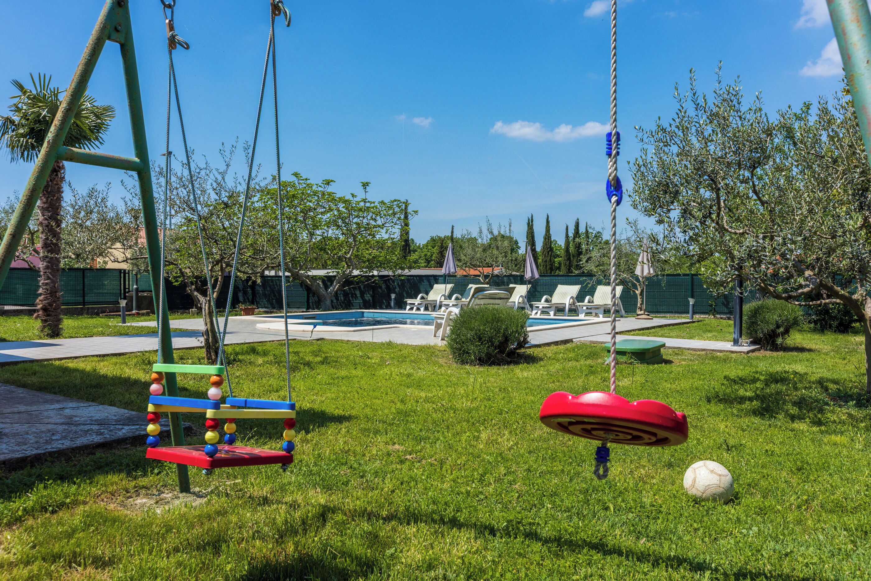 Un parque infantil con columpios y un balón, una piscina con sillas de descanso, y una valla verde.