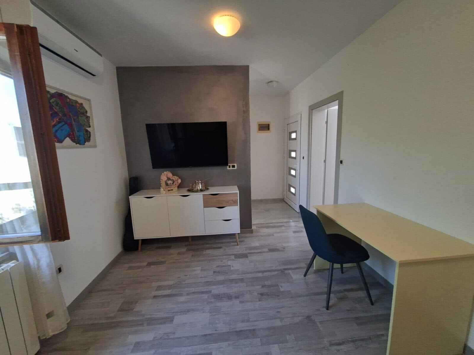 A room with a flat-screen TV mounted on the wall, a white cabinet with drawers, a blue chair, and a radiator on the left.