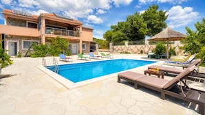 Ein Schwimmbecken mit Liegestühlen, eine Steinterrasse, ein Gebäude mit Balkon und ein Reetdachschirm.