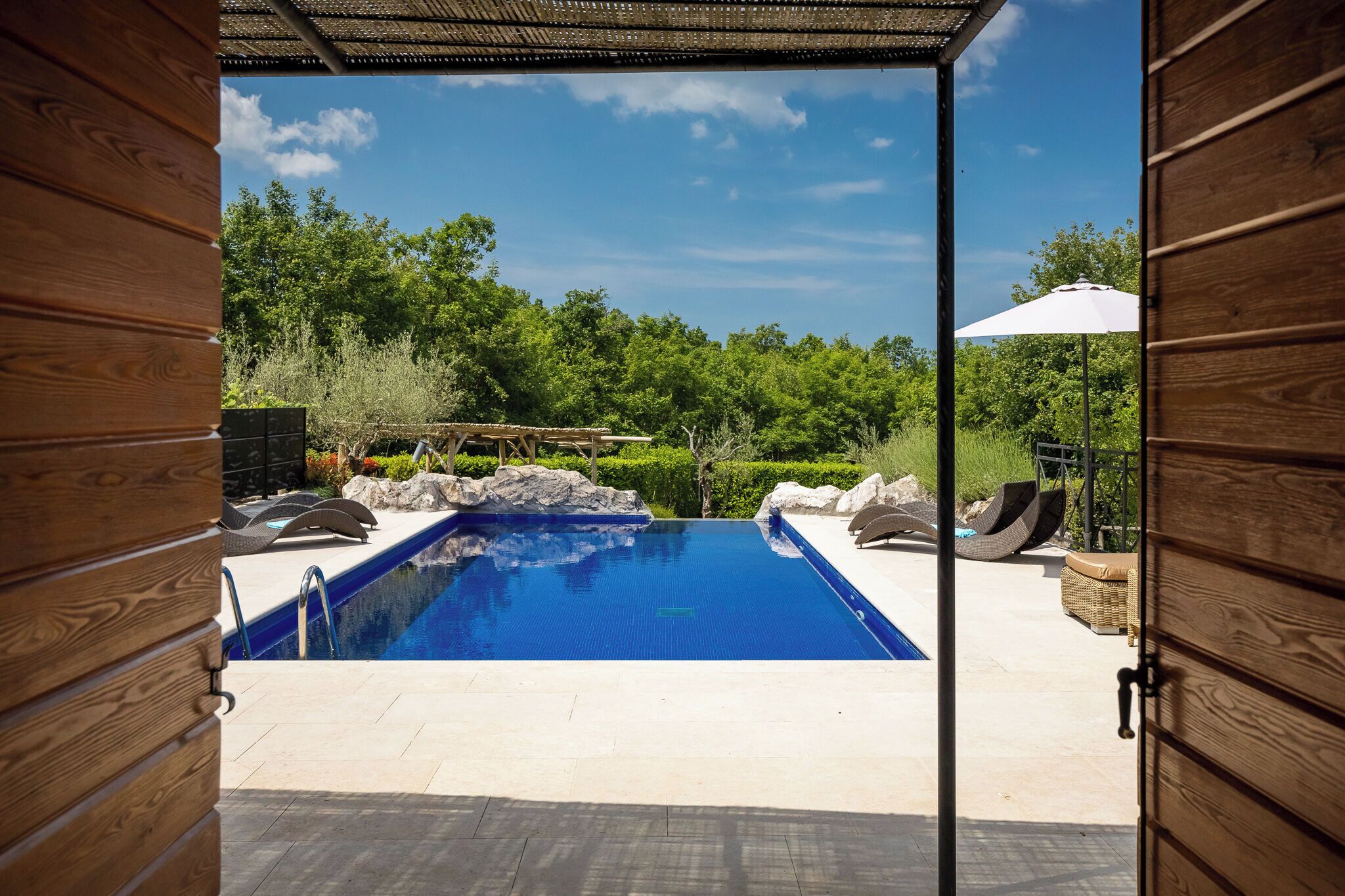 An outdoor swimming pool with a view of a forested area, surrounded by wooden walls and a pergola.