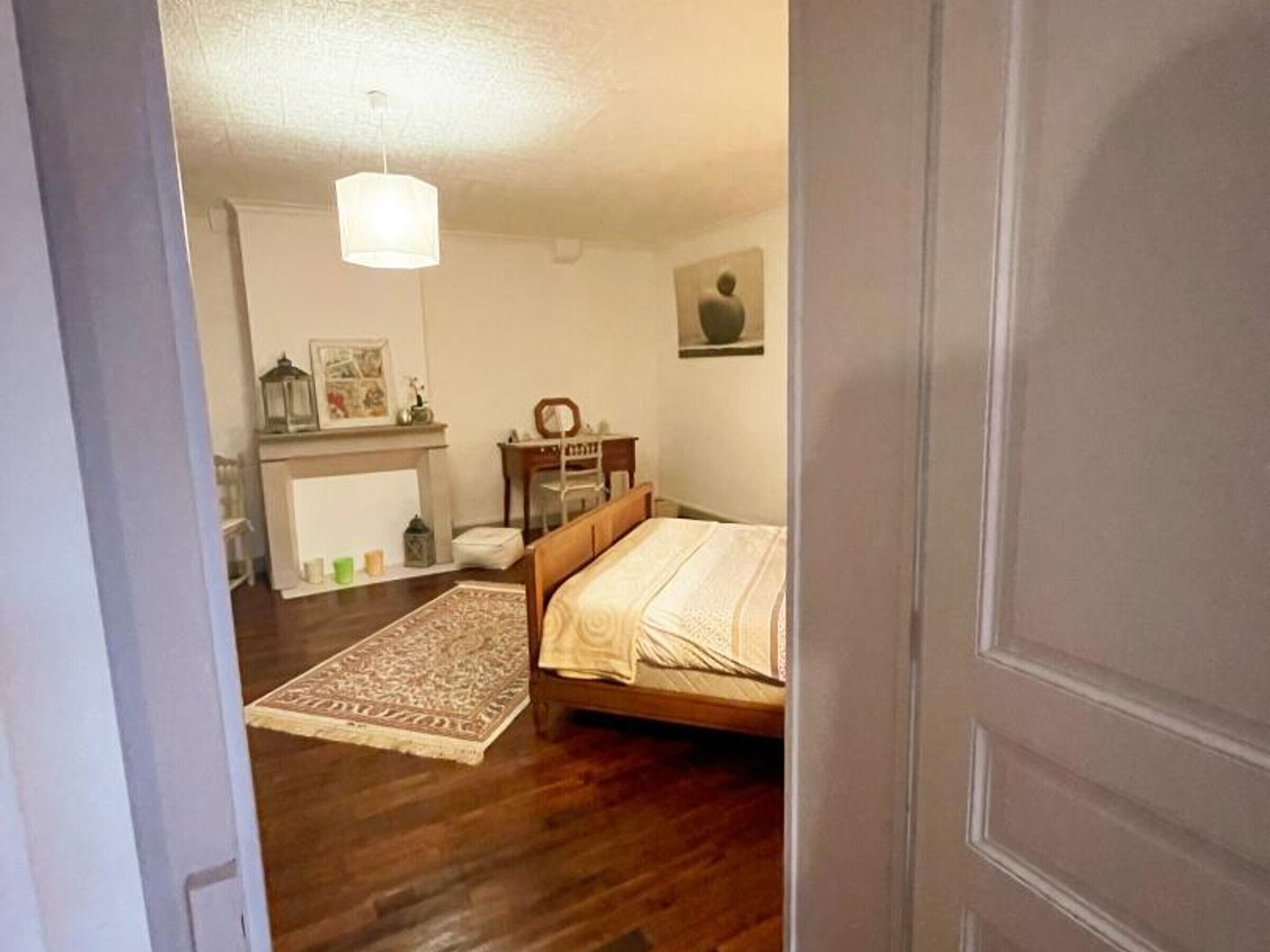 A bedroom with a wooden bed, a nightstand, a dresser, a chair, a rug, a hanging light fixture, and a fireplace.