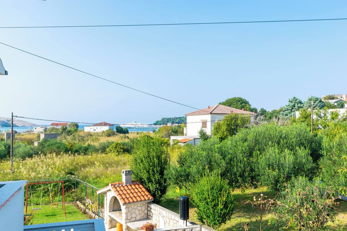 A residential area with a stone barbecue structure, a swing set, and a view of the sea and distant buildings.