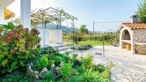 Un giardino con un vialetto in pietra, un pergolato con piante rampicanti e una costruzione in pietra con un camino.