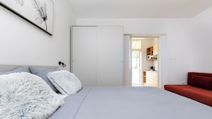 Una camera da letto con un letto, un divano rosso, un tappeto bianco e soffice, e una porta bianca che dà su una cucina.