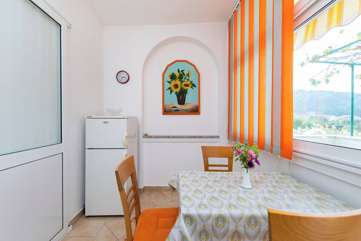 A small room with a dining table, chairs, a refrigerator, a clock, and a window with orange curtains.