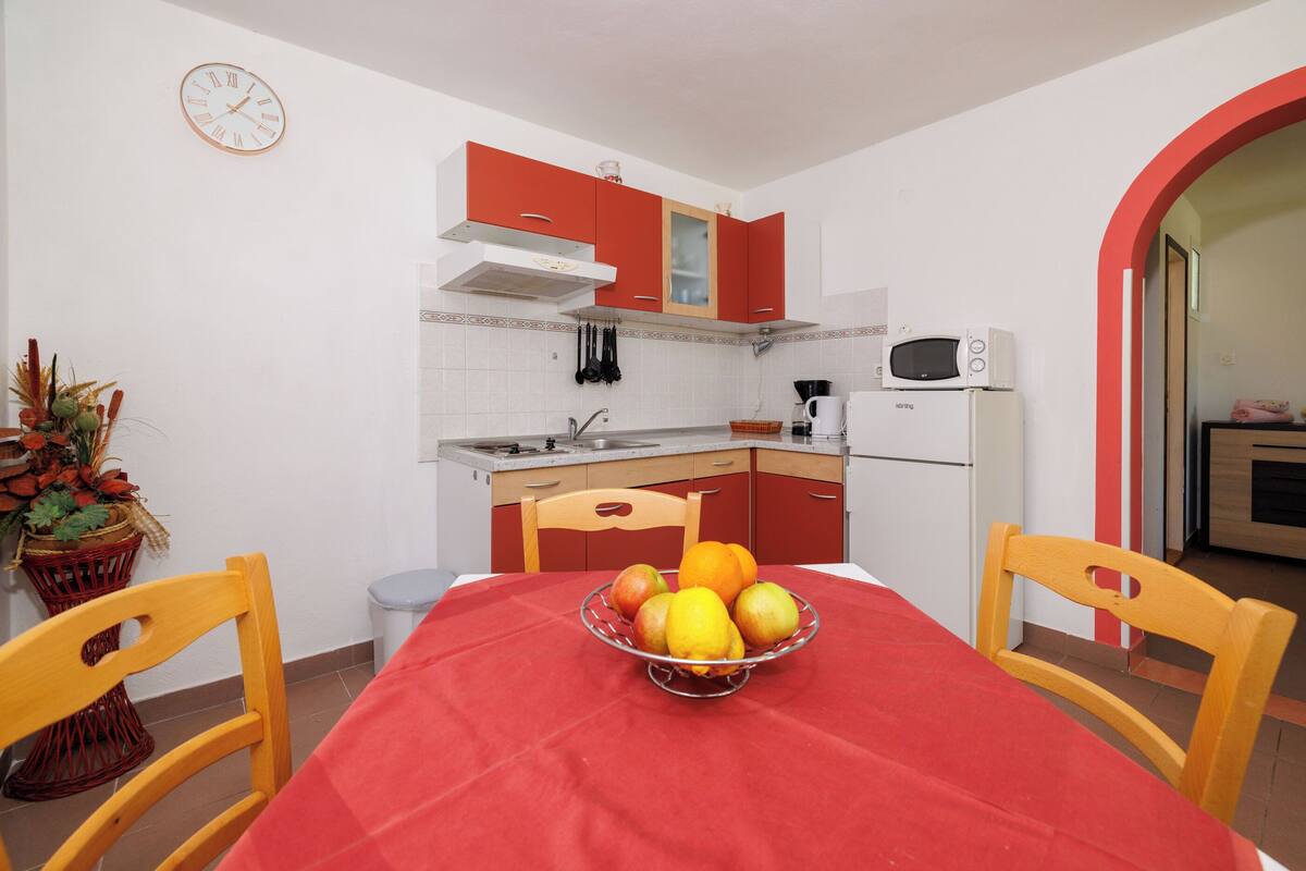 A compact kitchen with red cabinets, a microwave, and a sink. A fruit bowl is on the table, and there's a clock on the wall.