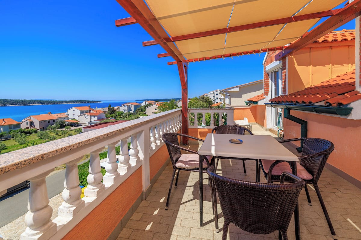 A balcony with wicker chairs and a table, overlooking a coastal view with houses and the sea.