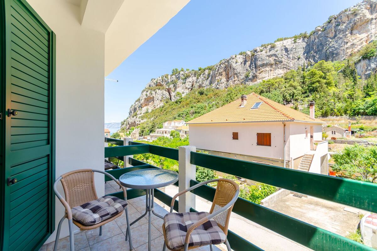 A balcony with two wicker chairs and a small table, overlooking a mountainous landscape with buildings.