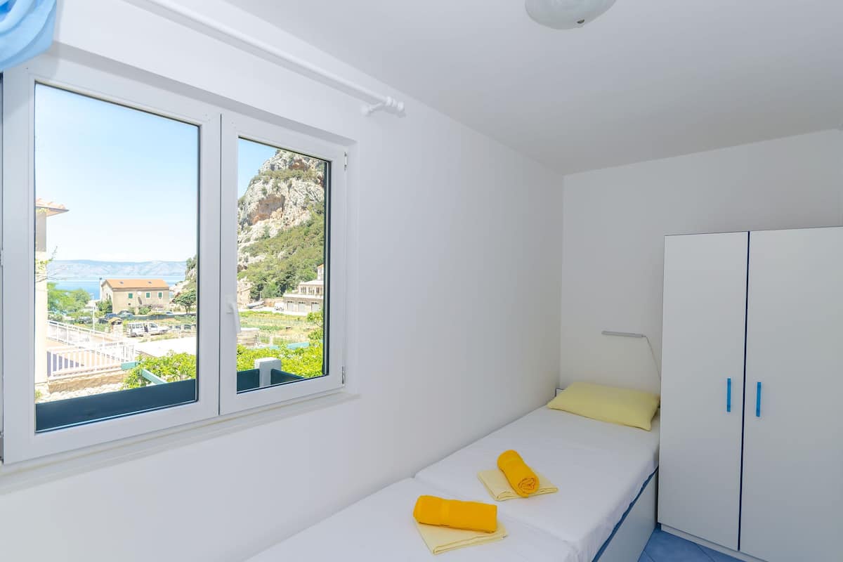 A small, bright room with a bed, a wardrobe, and a window with a view of buildings and greenery.