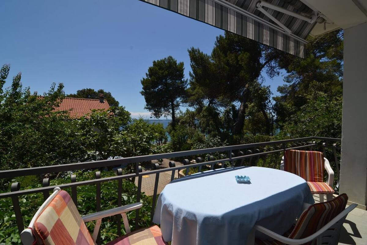 Ein Balkon mit Tisch und Stühlen, der auf eine Küstenlandschaft mit Bäumen und einem Gebäude mit rotem Dach blickt.