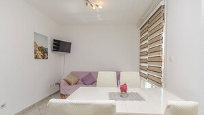 A room with a white dining table, a sofa with cushions, a TV mounted on the wall, and a window with blinds. - Apartments Jopa (Vodice)
