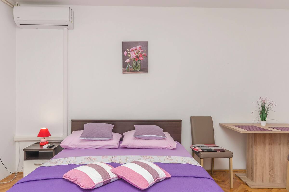 A neatly arranged bedroom with a bed, bedside table, lamp, and a small desk with a plant.