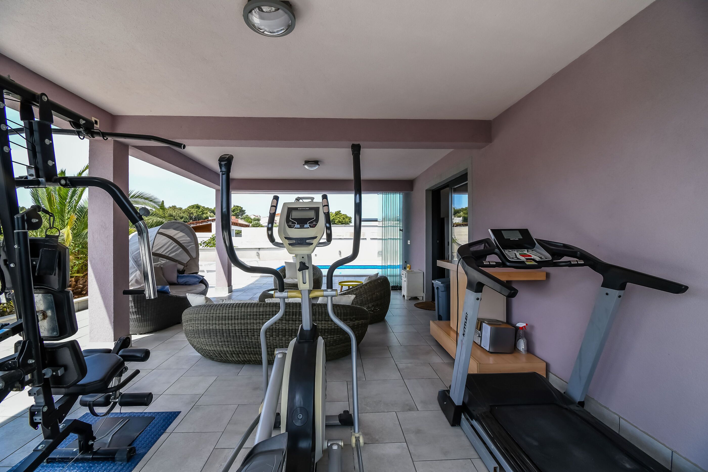 A home gym with exercise equipment, including a treadmill, elliptical, and weight machine, with a view of the outdoors.