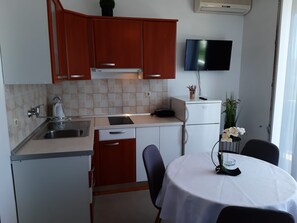 A compact kitchen with a sink, stove, and a small refrigerator. There's a dining table with chairs and a TV mounted on the wall. - Apartments and Room Kate (Tisno)
