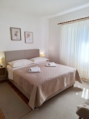 A neatly made bed with a beige bedspread and pillows, two bedside lamps, a framed picture on the wall, and a large window with sheer curtains. - Apartments and Room Kate (Tisno)
