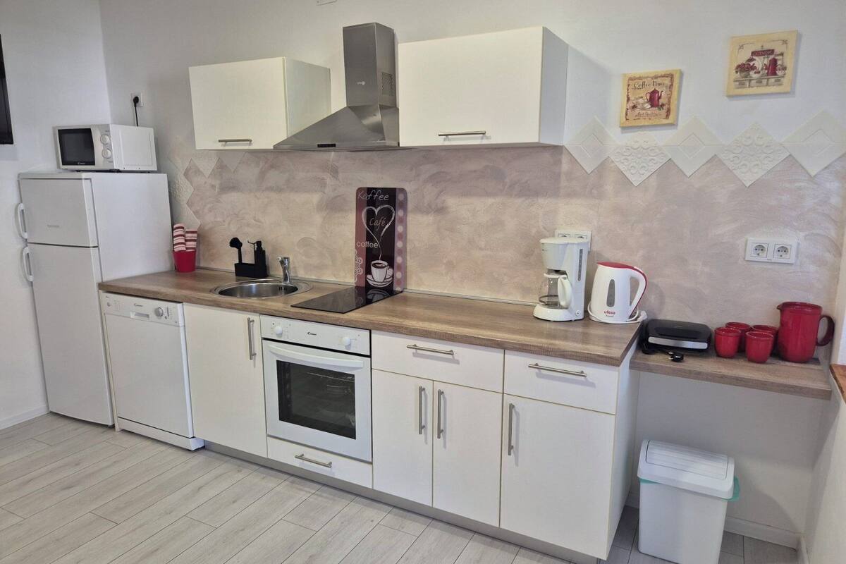 A modern kitchen with white cabinets, a built-in oven, a microwave, a coffee maker, and a trash bin.