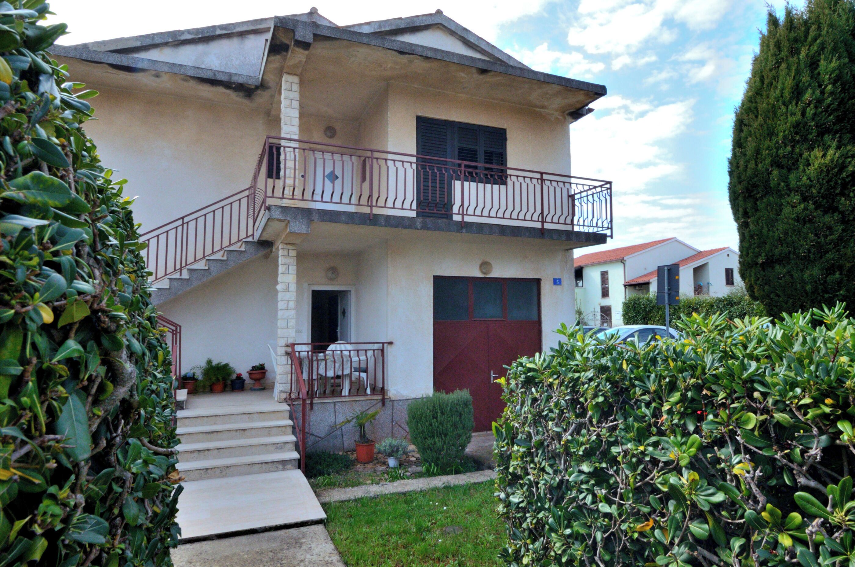 A two-story building with a balcony, a red door, and a small garden area with potted plants.