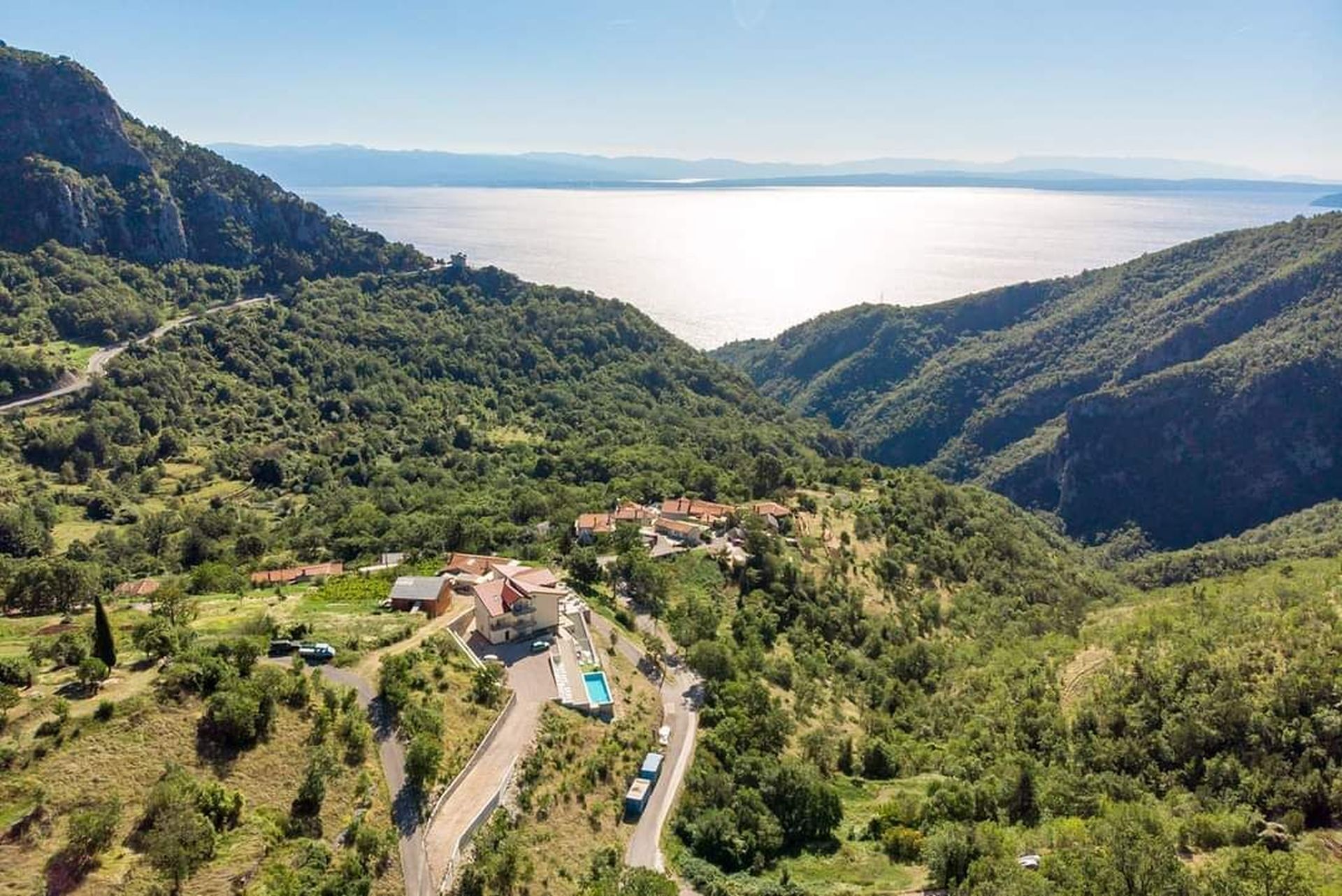 A mountainous landscape with a valley, a road, and a cluster of buildings with a swimming pool.