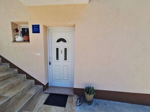 Une porte blanche avec une plaque portant un numéro, une petite fenêtre ornée d’un pot de fleurs et un escalier menant à l’entrée.