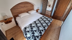 Un dormitorio con cabecera de madera, una cama con ropa de cama blanca, una colcha de flores en blanco y negro, una mesita de noche con una lámpara y un espejo en la pared.