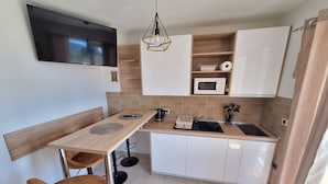 Cocina compacta con televisor, microondas y lavabo. Hay dos taburetes y una lámpara colgante.