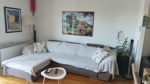 A cozy living room with a sectional sofa, a small round table, and a radiator. There are two framed artworks on the wall and a potted plant on the right. - Apartments Ivo (Makarska)
