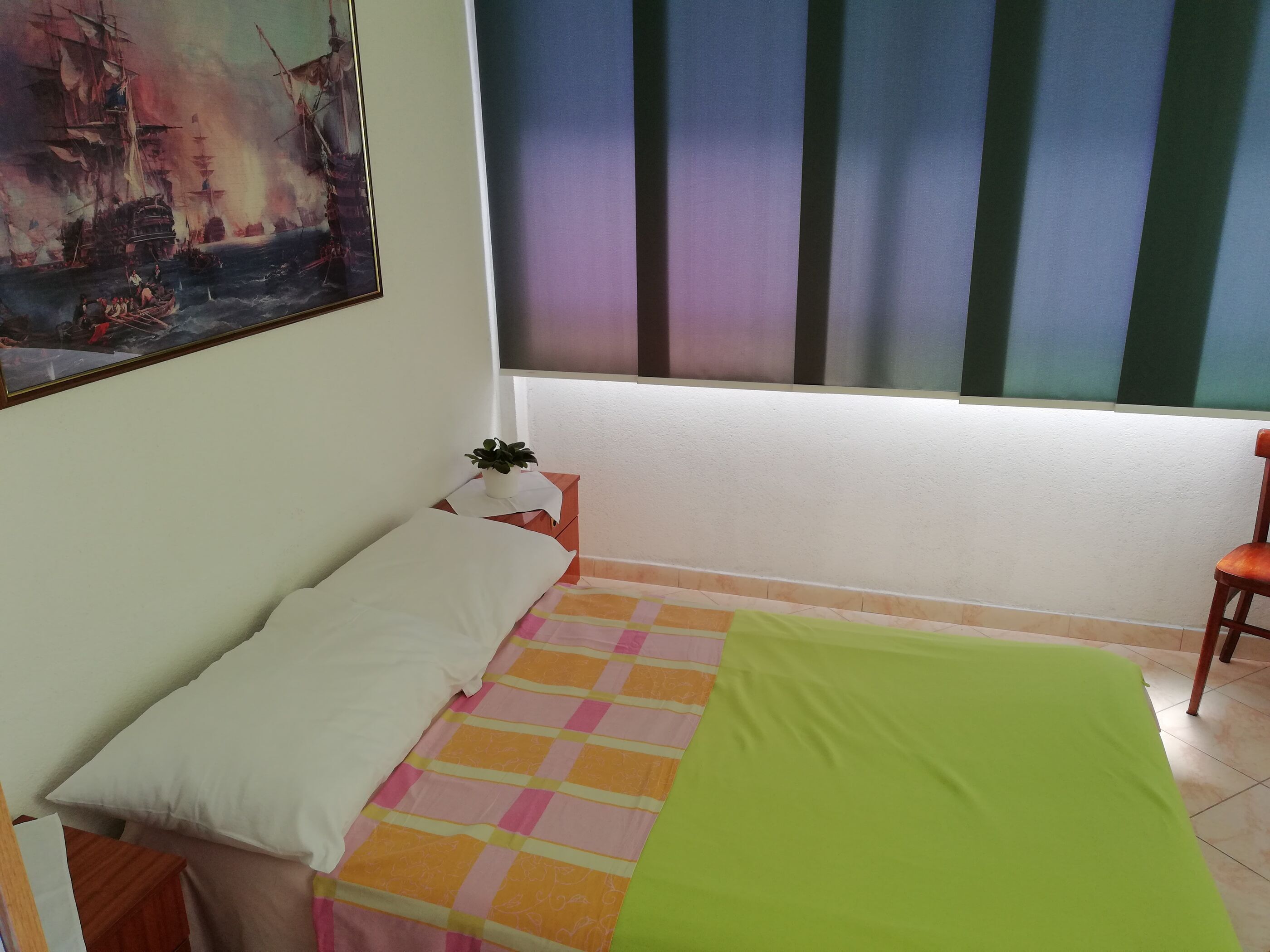Una stanza con un letto, un tavolino con una pianta, una finestra con persiane e un quadro appeso al muro.