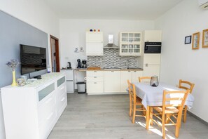 A compact kitchen with white cabinetry, a tiled backsplash, a built-in oven, and a dining area with a table and chairs. - Apartment Orlic (Senj)