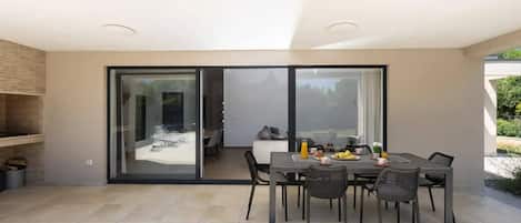 Uma área moderna para refeições ao ar livre, com mesa posta para dois, cercada por cadeiras, e uma porta de vidro deslizante que dá para um cômodo adjacente.