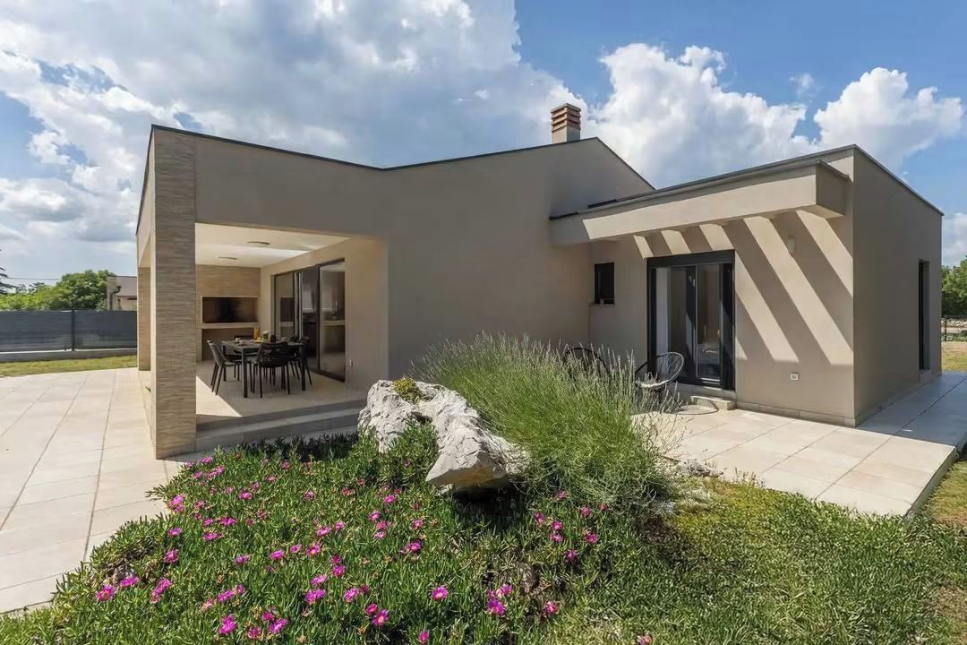 A modern two-story house with a patio area, a dining space, and a garden with pink flowers.