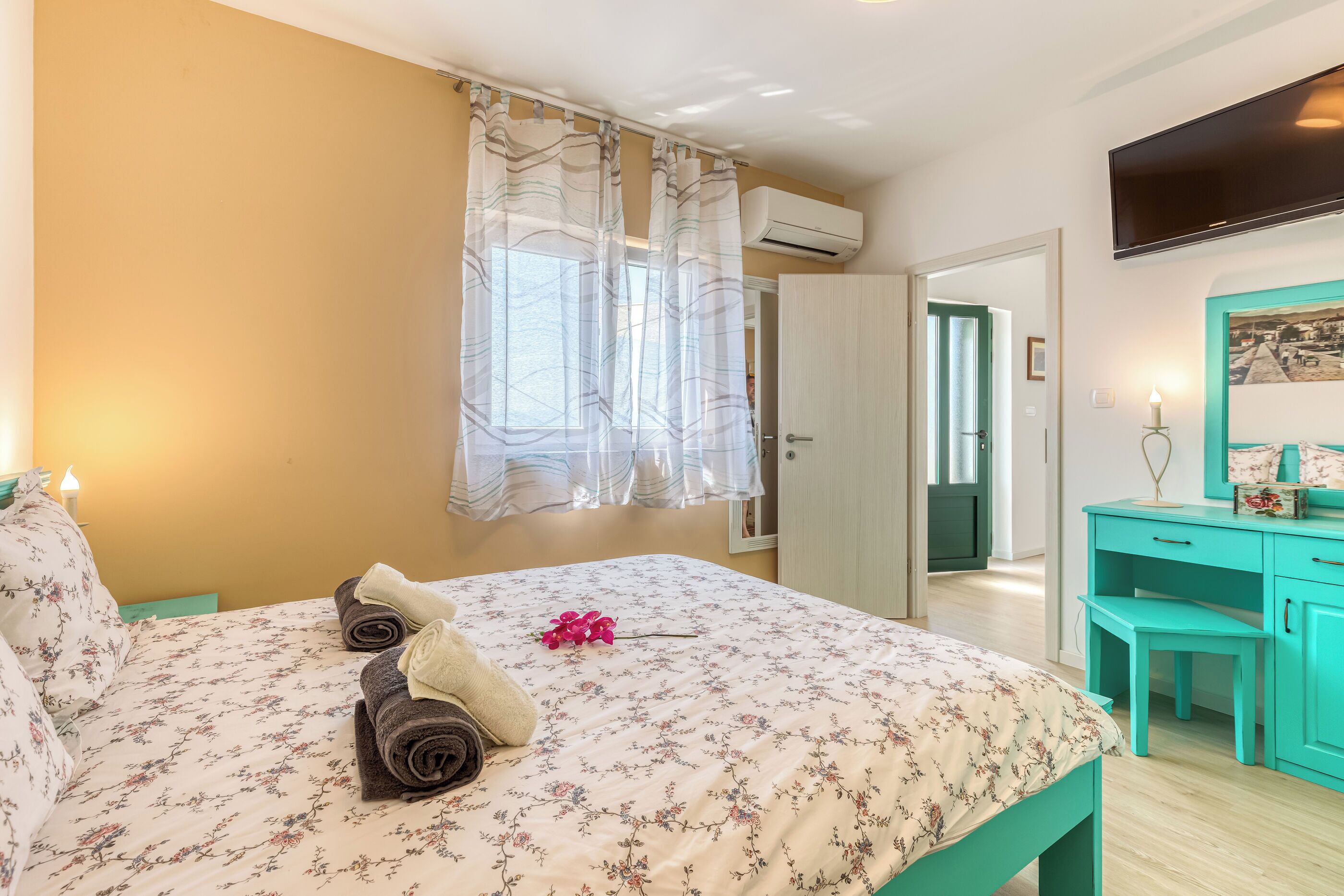 Une chambre avec une couvre-lit à motifs floraux, une paire de serviettes, une fleur rose, un bureau turquoise et un téléviseur fixé au mur.