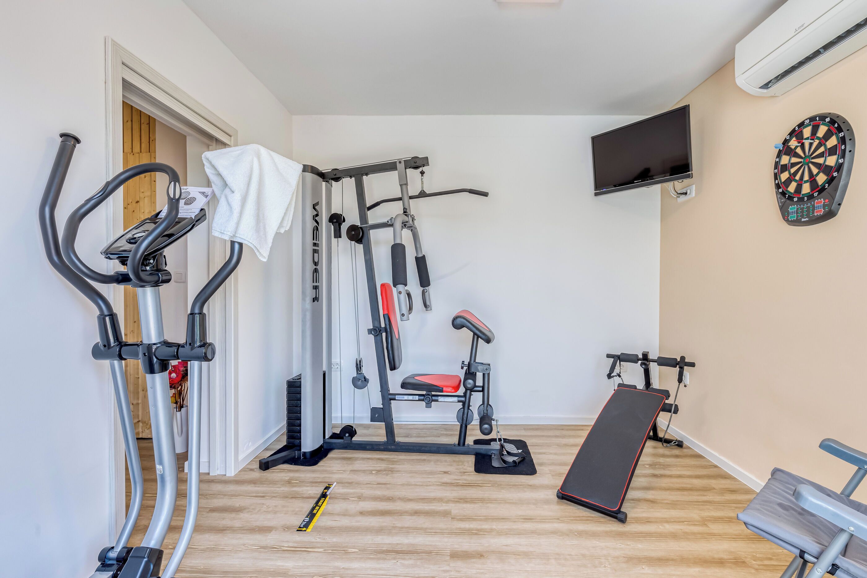 A home gym with an elliptical, a bench press, and a wall-mounted TV.