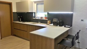 Une cuisine moderne avec des meubles en bois, un plan de travail blanc, un four encastré, une machine à café et une fenêtre équipée de stores.