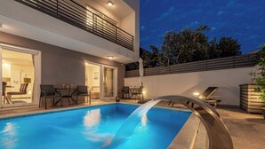 Una piscina moderna al aire libre con cascada, rodeada de sillones de descanso y una zona de comedor.