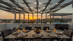 Un área de comedor en la azotea con mesas dispuestas para seis personas, con vista a un paisaje urbano al atardecer.