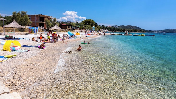 Una playa con aguas transparentes y poco profundas, sombrillas y gente relajándose.