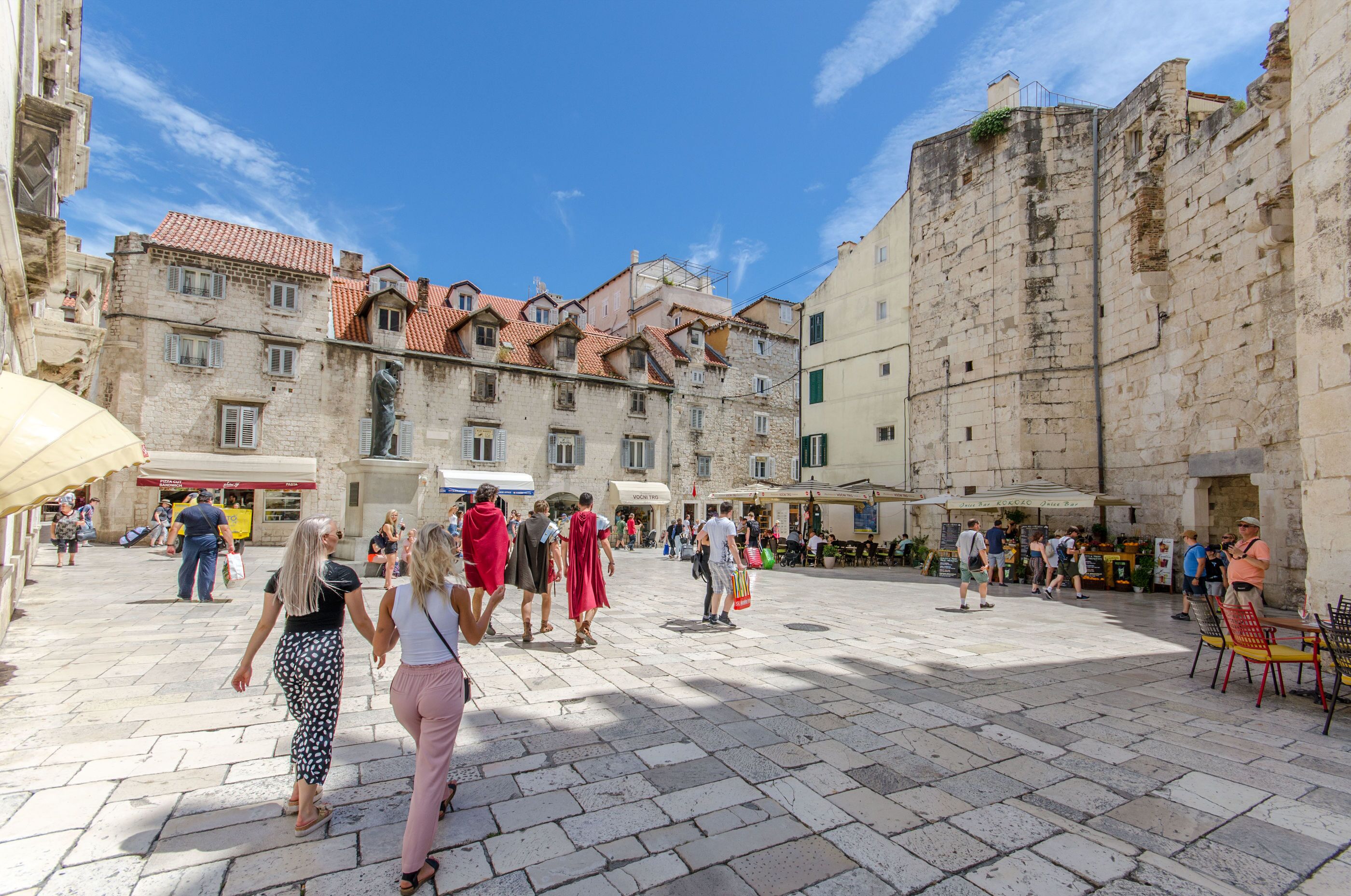 A historic square with stone buildings, outdoor seating, and people walking.