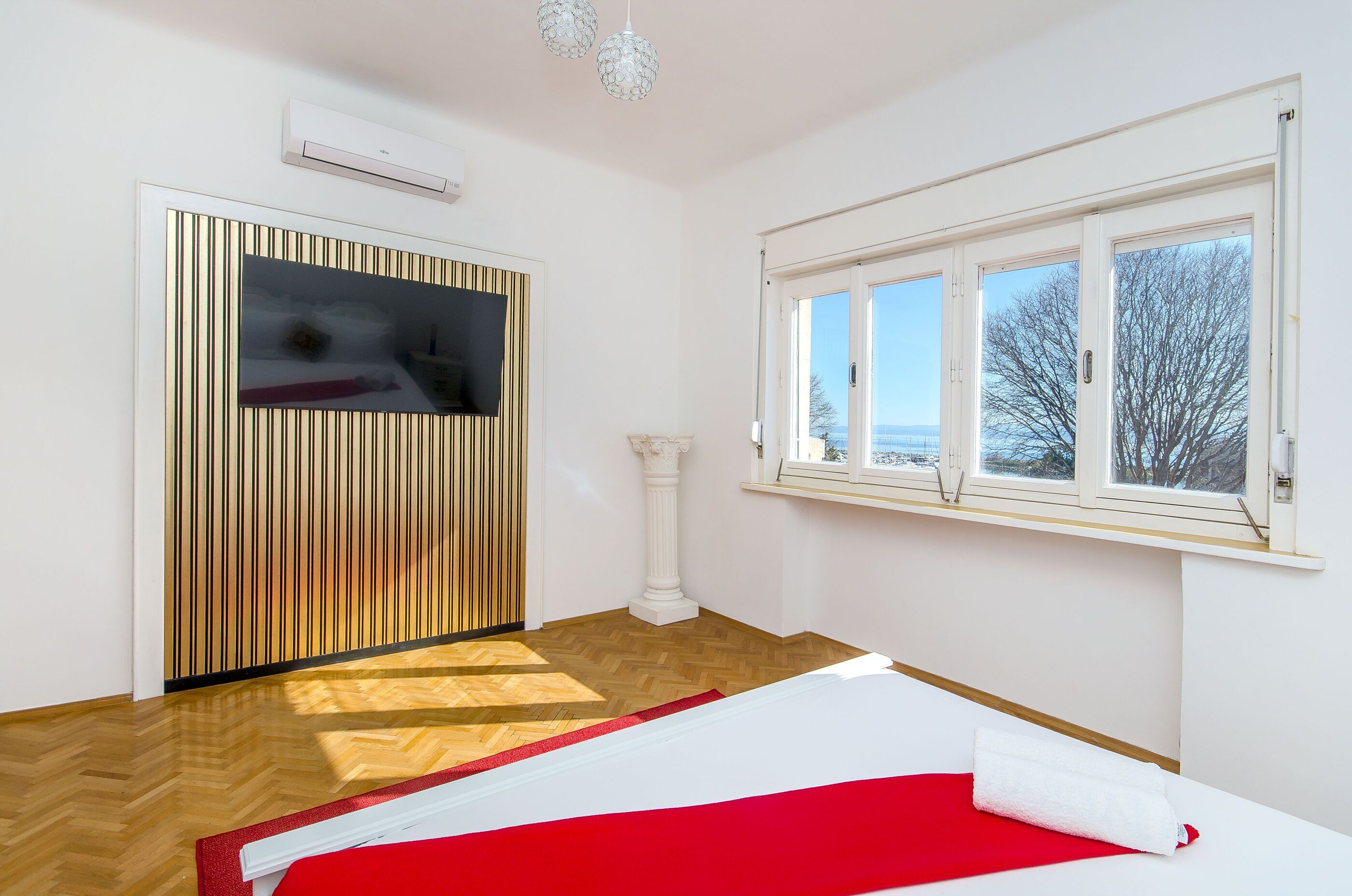 A room with a large window, a wooden floor, a white wall, and a red and white bedspread.