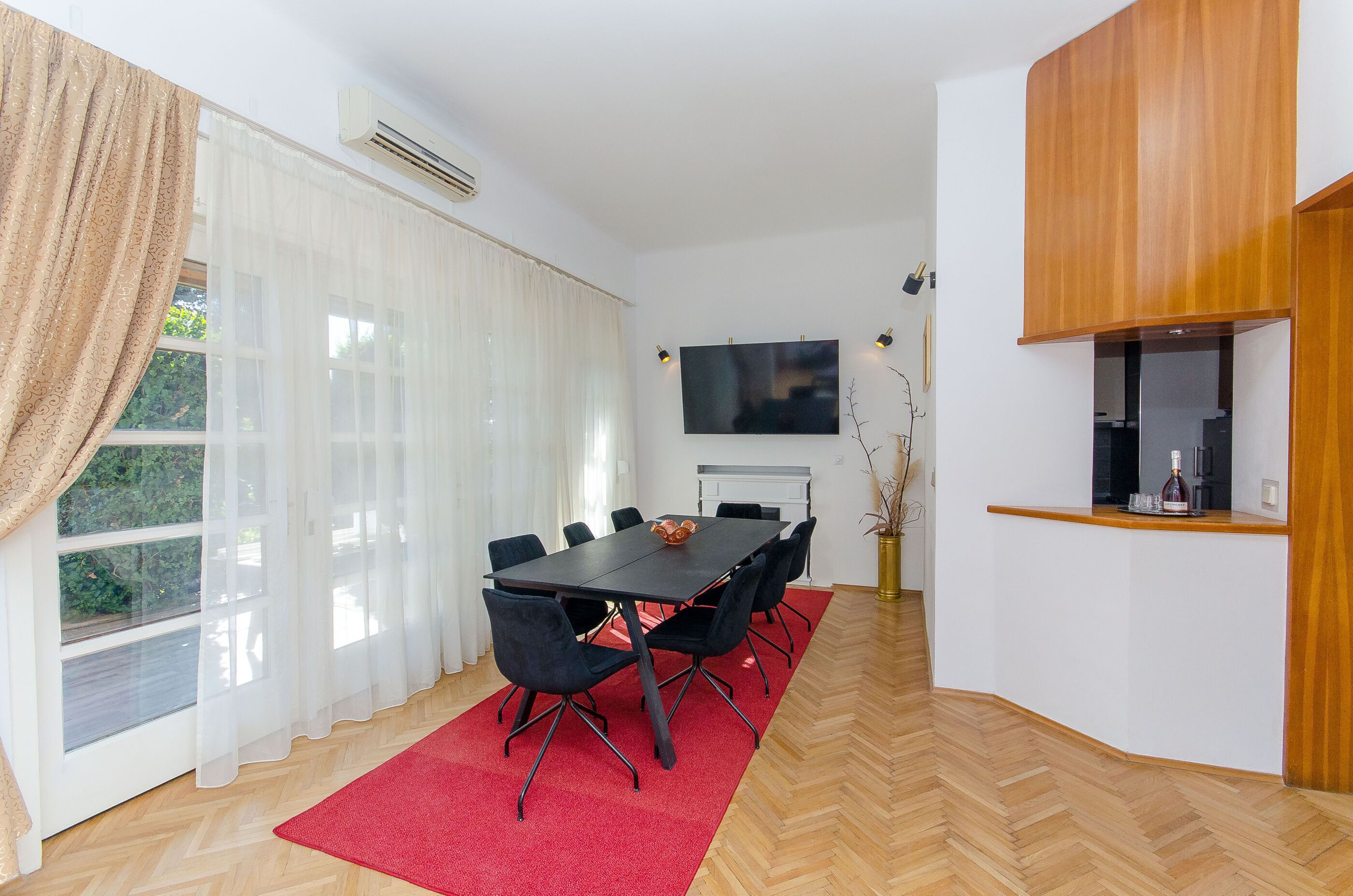 A modern room with a large dining table, black chairs, a red rug, a TV mounted on the wall, and a wooden cabinet.