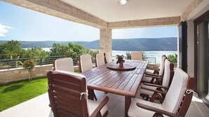 A covered outdoor dining area with a wooden table and chairs, a view of the sea, and a stone wall. - Villa Royal Blue (Rasa)