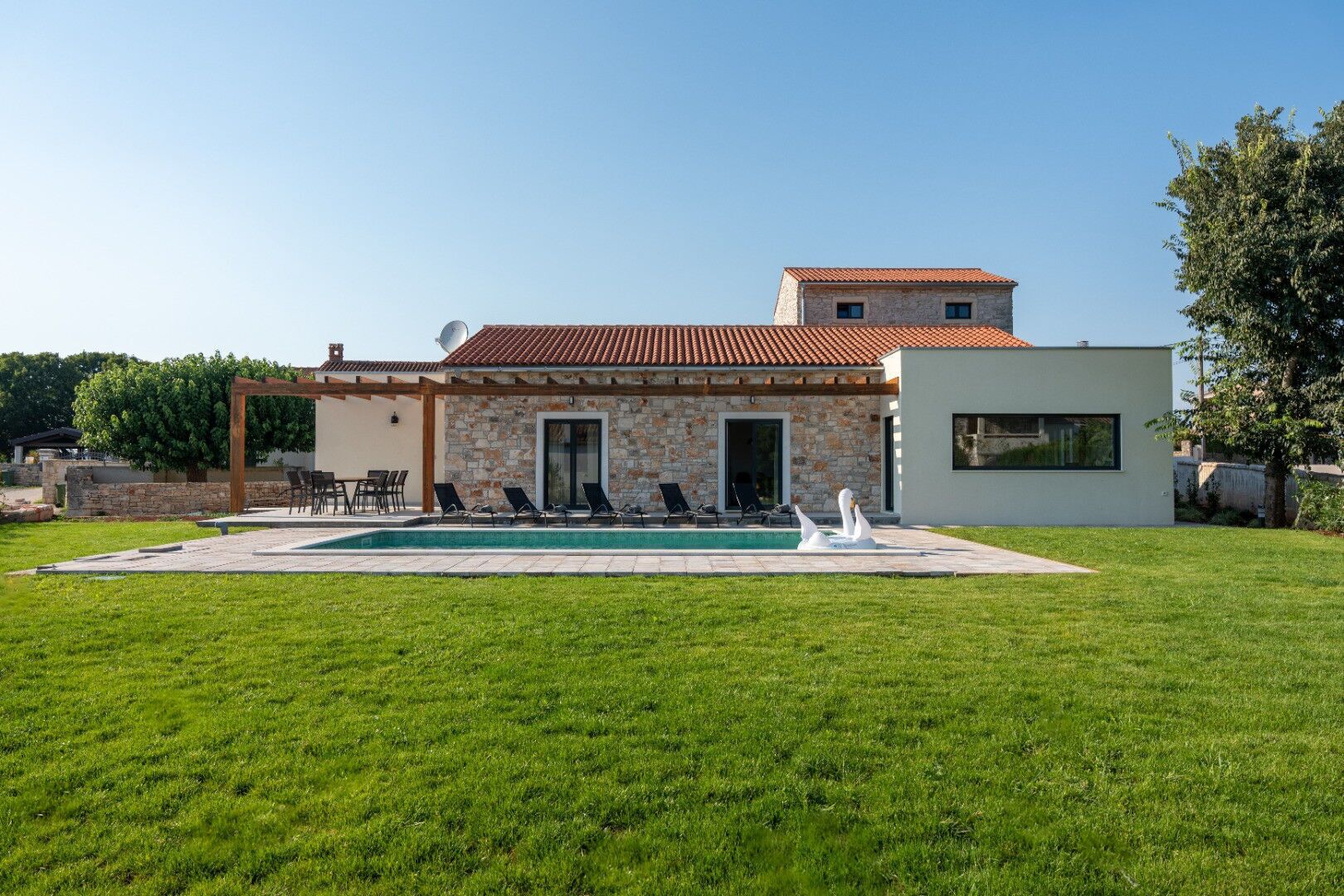 Una casa de dues plantes amb façana de pedra, una piscina amb gandules i una pèrgola.