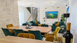 A modern living room with a wooden dining table, green and yellow chairs, a staircase, and a large TV screen displaying a scenic view. - Villa Nikoma (Dicmo)