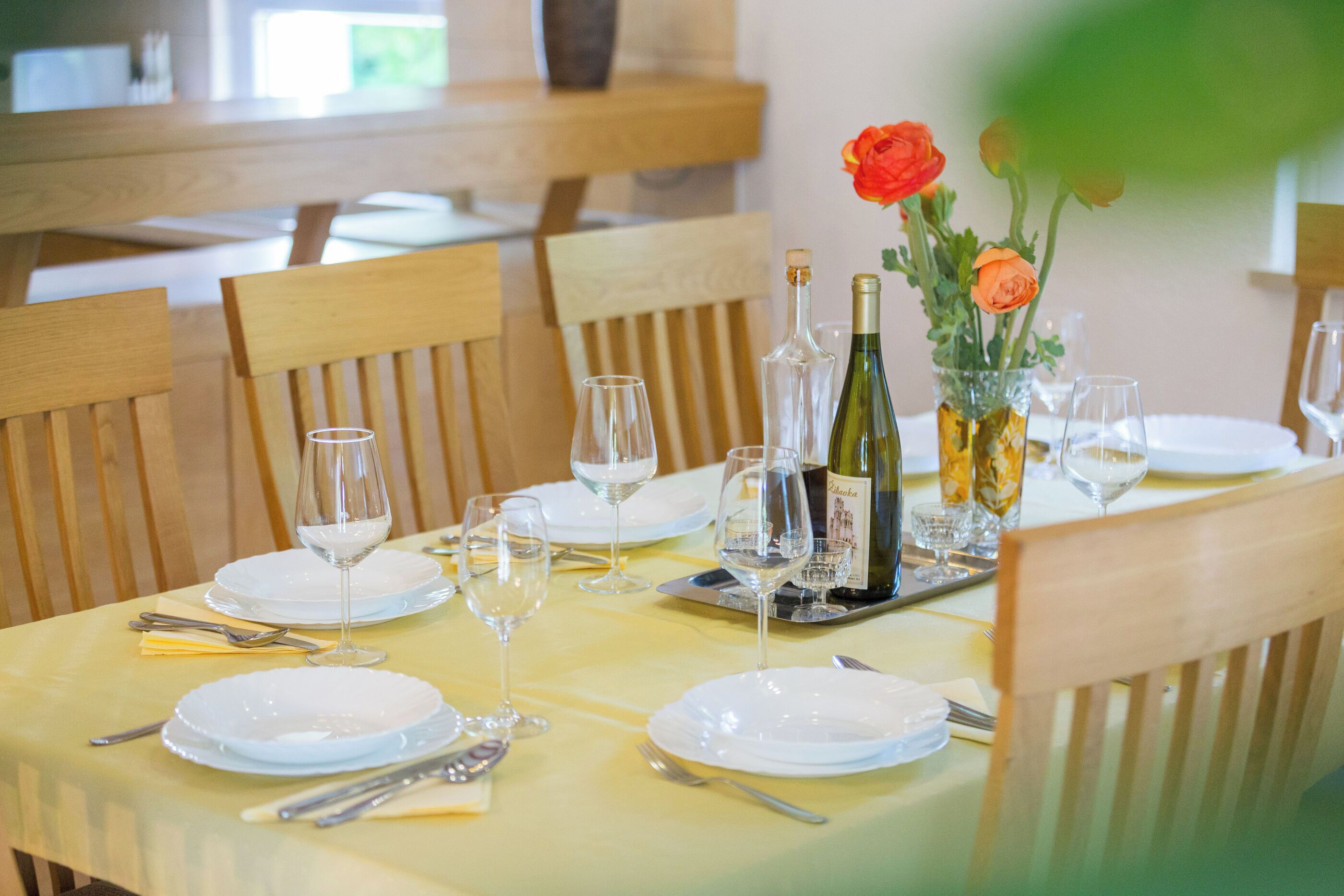 A dining table set with white plates, wine glasses, and a bottle of wine, surrounded by wooden chairs.