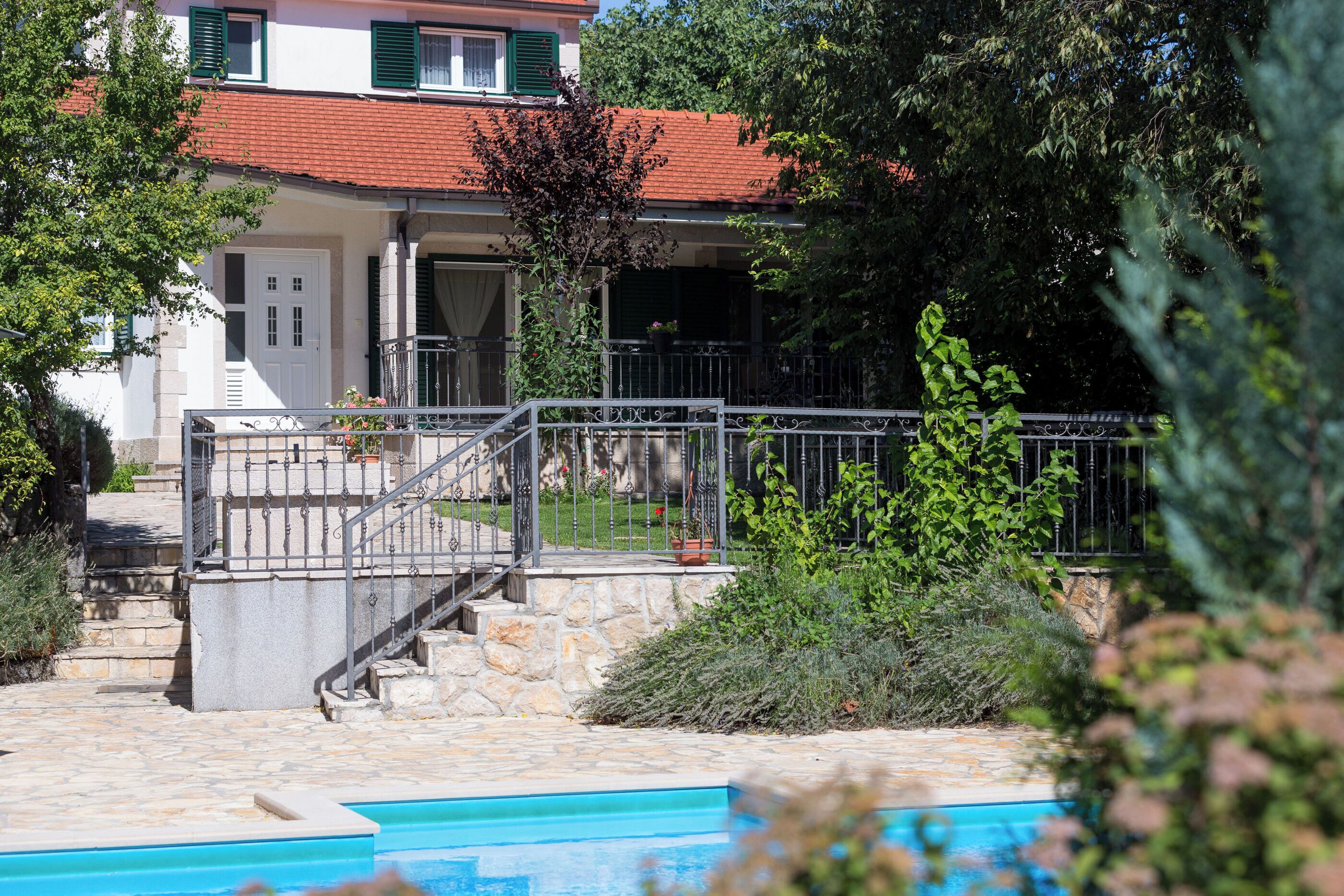 A two-story house with a stone staircase leading to a balcony, surrounded by greenery and a swimming pool.