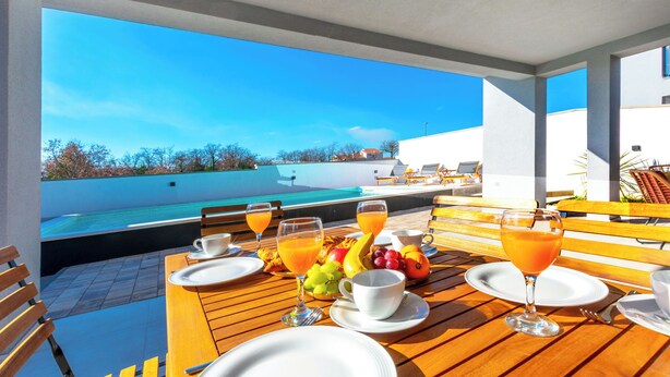 Una mesa de desayuno con platos, vasos y fruta, con vista a una piscina y un cielo azul despejado.