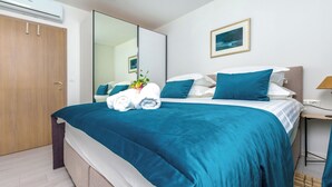 Una cama bien hecha con un edredón verde azulado, una almohada blanca y una almohada azul. A la izquierda hay un armario de madera y, a la derecha, una mesita de noche con una lámpara.