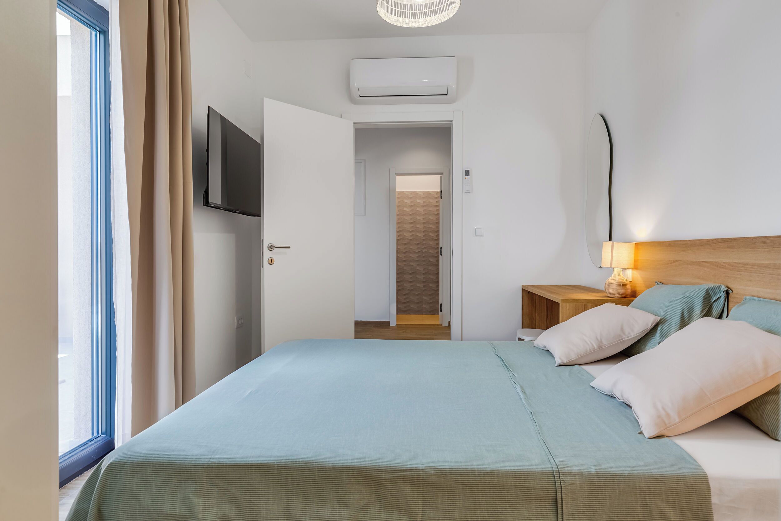 A bedroom with a bed, a TV, an air conditioner, a nightstand with a lamp, and a view of a hallway.