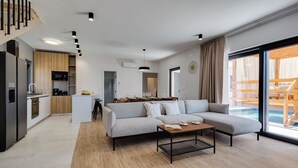 A modern living room with a grey sofa, a coffee table, and a dining area with a white table and chairs. There is a kitchen with a built-in oven and a dining table. The room has large windows with a view of a pool. - Villa A Lauda (Pakostane)
