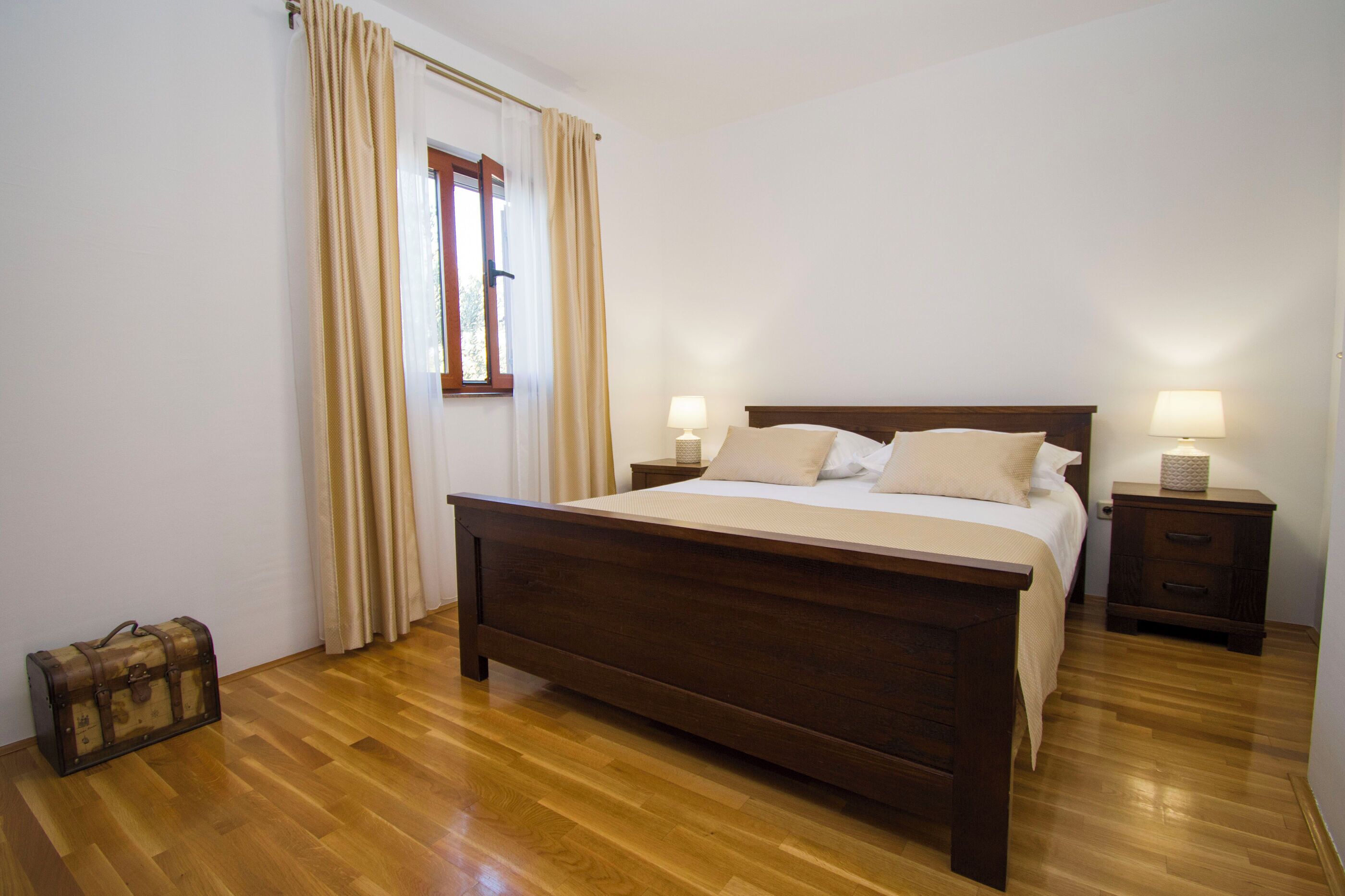 Un dormitorio con cama de madera, dos mesitas de noche con lámparas, una ventana con cortinas y una maleta en el suelo.