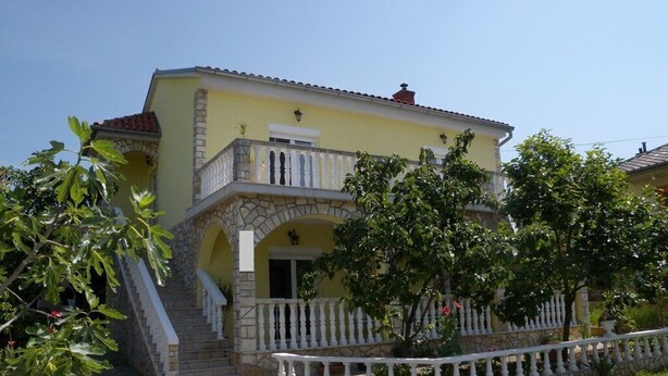Una casa a due piani con un balcone, una scala in pietra e un giardino con alberi e piante.